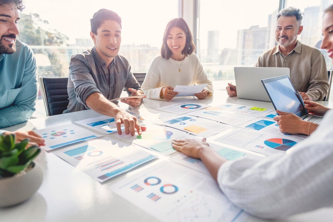 Business professionals around a table looking at printed out graphs and charts. Some are smiling and they are all collaborating with one another.
